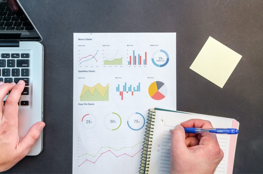 A photo of a laptop, a printout of various charts, and a person writing in a notebook.