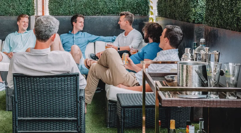 six men sitting outdoors conversing and laughing while holding cups of alcohol, bar cart on the bottom right side. photo cred: austin distel
