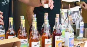 Hand passing mini paper sample cup, surrounded by various Two Drifters bottles on a table. Photo by Amy Vann.